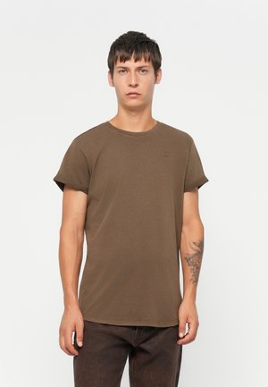 Jeune homme aux cheveux bruns courts, portant un t-shirt marron à manches retroussées et un pantalon sombre, se tenant devant un fond blanc uni.