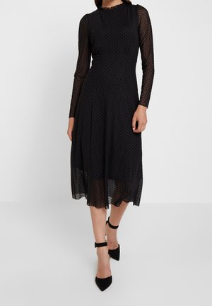 Kvinde iført en sort, gennemsigtig polkaprik midi-kjole med lange ærmer, kombineret med sorte ankelrem-spidse hæle.