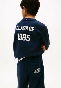 Junge in navyblauem Outfit mit "CLASS OF 1985" auf der Rückseite des Sweatshirts und weißem Logo auf der Tasche der Sweatpants, steht mit verschränkten Armen.