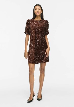 Le modèle se tient devant un fond blanc, vêtu d’une mini-robe marron foncé à sequins et à manches bouffantes, avec des talons noirs.