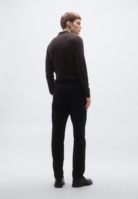 Rudas megztinis su apykakle, juodos kelnės ir juodi batai. Modelio nugara atsisukusi į kamerą prieš neutralią foną.