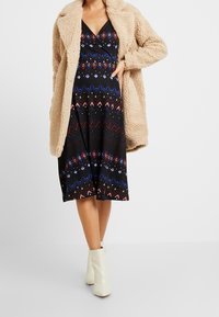 Černé šaty s barevnými geometrickými vzory, vrstvené pod opáleným huňatým kabátem. Mají výstřih do V a střední délku, doplněné bílými kotníkovými botami.