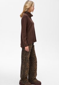 Kobieta stojąca z boku, ubrana w ciemno-brązowy sweter z golfem, luźne spodnie w panterkę i brązowe buty na platformie, na białym tle.