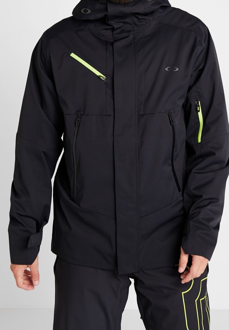 Veste noire avec des zippers vert fluo sur la poitrine et les manches, deux poches zippées noires à l'avant, portée avec un pantalon noir ornés d'un design vert fluo.