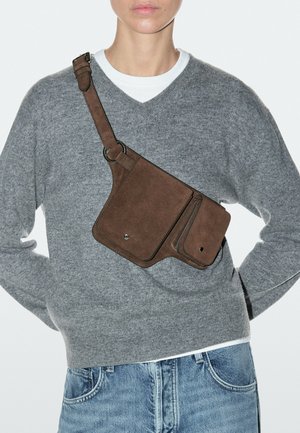 Persoon met een grijze trui met V-hals, wit overhemd, blauwe spijkerbroek en een bruine suède schoudertas met twee voorzakken die zijn vastgemaakt.