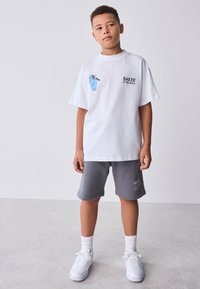 Berniukas dėvi baltą per didelį marškinėlį su grafiškais raktų paveikslėliais ir užrašu „BAKER“, pilkus šortus, baltas kojines ir baltus sportinius batelius, stovintis ant betono grindų.