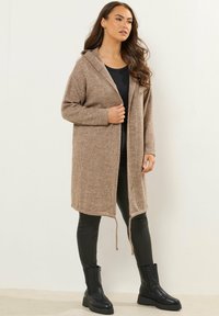 Larga chaqueta de punto beige con capucha, textura acanalada, de frente abierto y lazos ajustables. Combinada con leggings negros y botas negras gruesas.