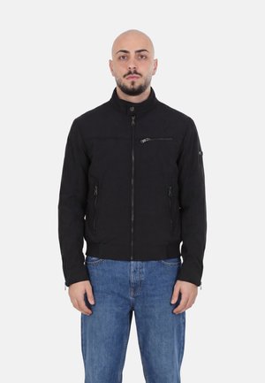 Uomo con testa rasata che indossa una giacca nera con zip e tre tasche frontali con zip e jeans blu, in piedi davanti a uno sfondo uniforme.