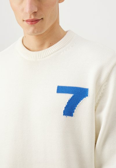 Pull en tricot blanc avec un grand chiffre 7 bleu sur la poitrine. Le tissu semble doux avec un col côtelé texturé.