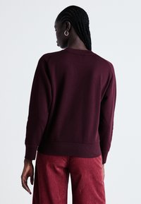 Femme aux cheveux tressés portant des boucles d'oreilles en argent, un sweat à capuche bordeaux foncé et un pantalon en velours côtelé rouge, se tenant devant un fond uni.