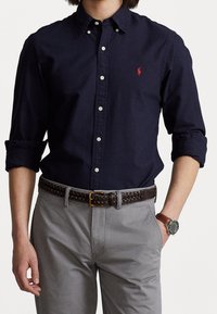 Hombre vestido con una camisa de botones azul marino con logo rojo, pantalones grises, cinturón trenzado marrón y reloj de pulsera marrón con cara verde, con las manos en los bolsillos.