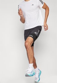 Weißes Sportshirt mit schwarzen Streifen, schwarze Shorts mit grauen Akzenten und hellblaue Laufschuhe mit lila Details.