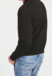 Sudadera negra con cuello redondo y puños de canalé, combinada con jeans de mezclilla azul claro que tienen bolsillos traseros y detalles de costura.