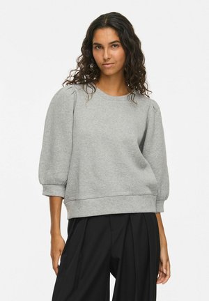 Donna con capelli ricci indossa una felpa grigia con maniche a sbuffo e pantaloni neri a gamba larga, in piedi davanti a uno sfondo chiaro e uniforme.