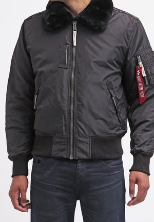 Personne portant une veste bomber sombre avec un col en fourrure, une fermeture éclair devant, deux poches latérales et une étiquette rouge "Remove Before Flight" sur la poche de la manche.