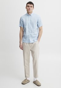 Camisa de lino de manga corta en azul claro con cuello abotonado, acompañada de pantalones beige y zapatos deslizables beige. Diseño simple y ligero.