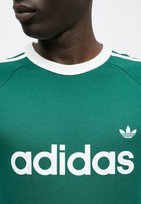 Πράσινο βαμβακερό μπλουζάκι με λευκή στρογγυλή λαιμόκοψη και τρεις λευκές ρίγες στους ώμους, με μεγάλο λευκό λογότυπο "adidas" και μικρό έμβλημα τριφυλλιού.