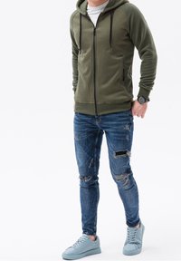 Felpa con cappuccio verde con cerniera e finitura testurizzata, jeans skinny blu usurati e sneakers azzurro chiaro, creando un outfit casual.