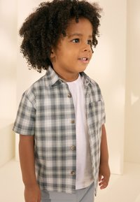Chemise à manches courtes à carreaux gris et blanc avec des boutons en bois, portée sur un t-shirt blanc. Enfant aux cheveux bouclés, expression souriante.
