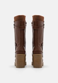Brown leather knee-high boots with a ribbed brown knit cuff, side zipper, and chunky textured heel. Decorative buckle and strap accents.
