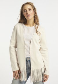 Beige fröcskölt kabát egyenes vonallal, puha anyagból készült, oldalzsebekkel, alatta egyszerű fehér blúzzal.