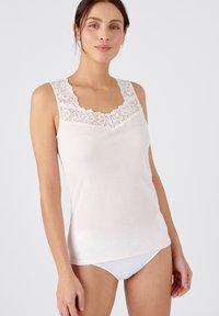 Top blanco sin mangas con escote de encaje floral, diseño ajustado y confeccionado en una tela suave y ligera. La modelo lleva un conjunto blanco.