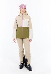 Beige en olijfgroene skijkacket met roze rits, gecombineerd met beige broeken. Het model draagt een roze beanie en zwarte skischoenen.
