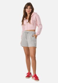 Kvinde stående med hånden i lommen iført pink cropped hættetrøje, grå shorts med snøre og røde sneakers mod hvid baggrund.