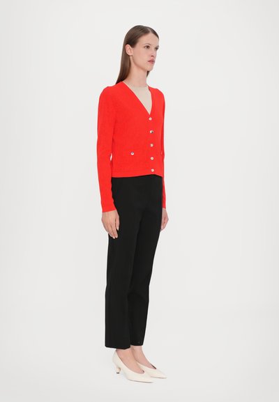 Cardigan lavorato a maglia rossa con scollatura a V, maniche lunghe e sei bottoni argentati. Indossato sopra una maglietta chiara, abbinato a pantaloni neri e tacchi beige.
