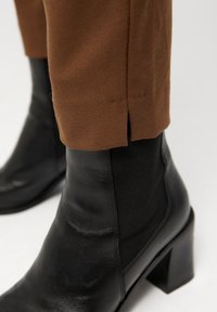 Svart läderankelboot med texturerad yta, med klossklack och elastiska sidopaneler, i kombination med bruna avsmalnande byxor.
