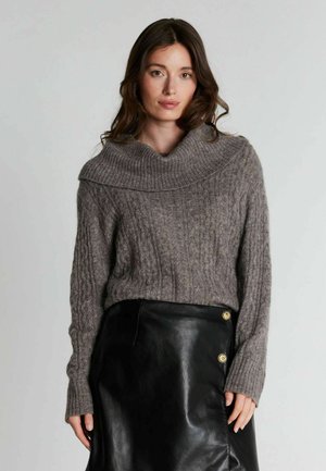 Femme aux longs cheveux bruns portant un pull en maille torsadée gris avec un col plié et une jupe en cuir noire avec des boutons dorés.