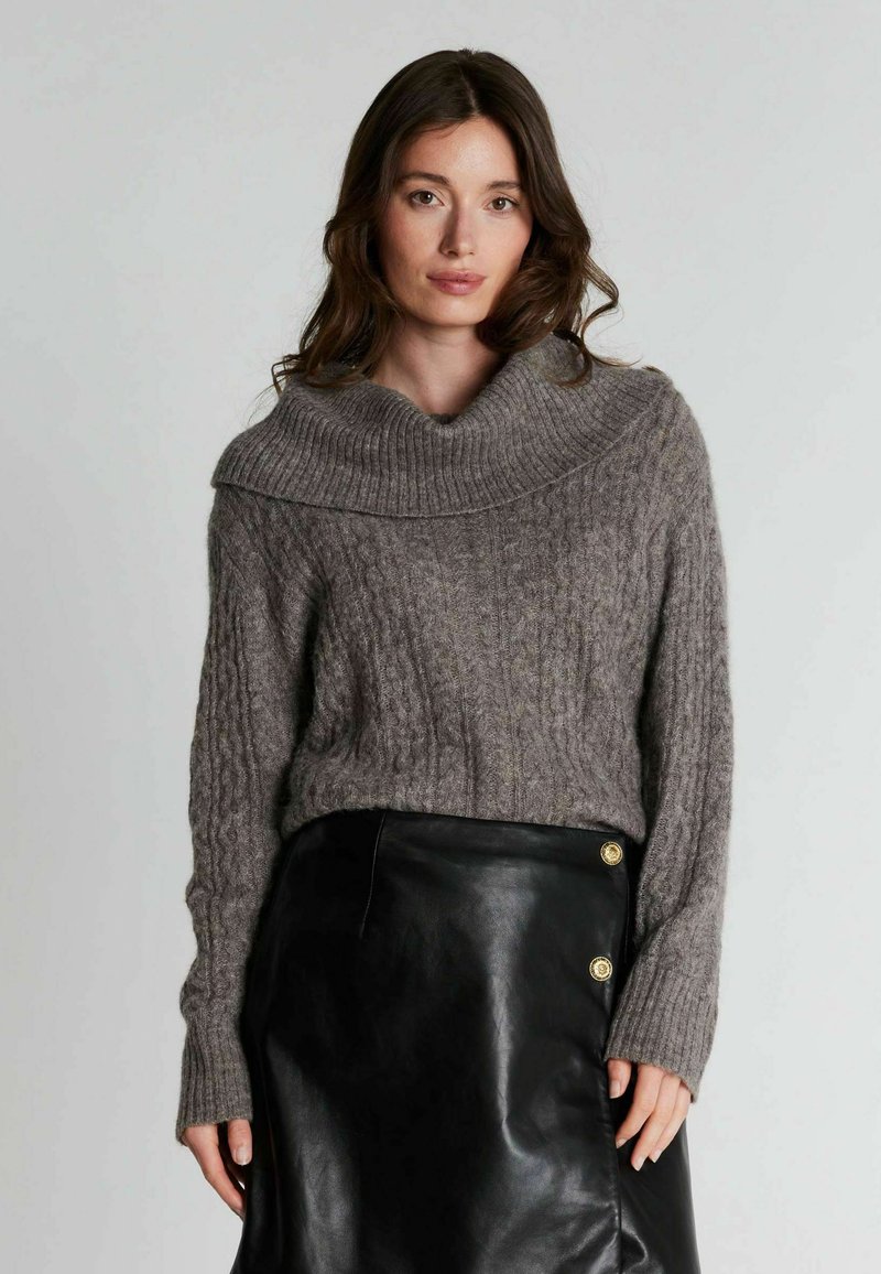 Femme aux longs cheveux bruns portant un pull en maille torsadée gris avec un col plié et une jupe en cuir noire avec des boutons dorés.