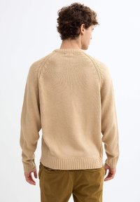 Suéter de punto beige con dobladillo y puños acanalados, que presenta una parte trasera texturizada y un cuello acanalado. Se lleva con pantalones caqui.