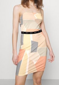 Kolorowa sukienka w geometryczne wzory, uszyta z miękkiego materiału, z regulowanymi ramiączkami spaghetti i markowym czarnym pasem w talii. Zawiera odcienie brzoskwini, szarości i beżu.