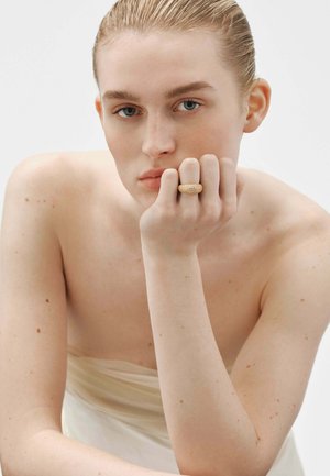 Anillo texturizado de color dorado llevado en una mano con los dedos apoyados en la cara; el fondo suave resalta los detalles de la joya.