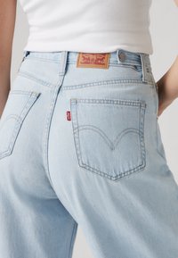 Light blue jeans featuring a high waist, curved back pockets, and a leather patch with a logo at the back, highlighting denim texture.