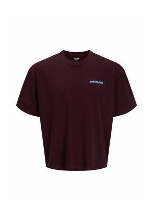 Teenage dreng iført en lyseblå kasket, mørkelilla t-shirt, mørke denimshorts, hvide sokker og beige sko, står og smiler på en ensfarvet baggrund.