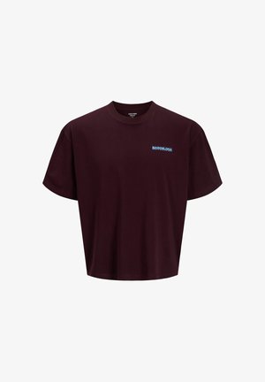 Teenage dreng iført en lyseblå kasket, mørkelilla t-shirt, mørke denimshorts, hvide sokker og beige sko, står og smiler på en ensfarvet baggrund.