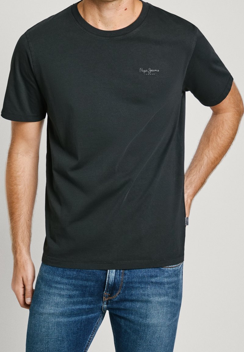 Uomo che indossa una maglietta nera con il logo di Pepe Jeans e jeans blu, in piedi con una mano in tasca su uno sfondo semplice.