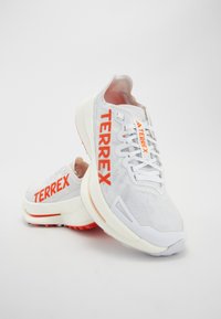 White athletic shoes featuring a mesh upper with orange "TERREX" branding, rubber sole, plush heel, and a streamlined design.
