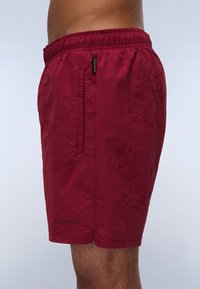 Rode shorts met een gestructureerd patroon, elastische tailleband en een zijzak met rits, met de merknaam "MANAFURI" aan de zoom.