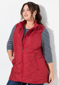 Rote Puffer-Weste mit Reißverschluss, hohem Kragen und zwei seitlichen Taschen. Sie hat ein dezentes geprägtes Muster und wird über einem schwarz-weißen gestreiften Oberteil getragen.