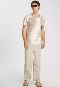 Pasek koszula z krótkim rękawem w kolorze beżowym i br ązowym, zestawiona z jasnobeżowymi spodniami. Nosi sandały i ciemne okulary przeciwsłoneczne. Widać tatuaż na ramieniu.