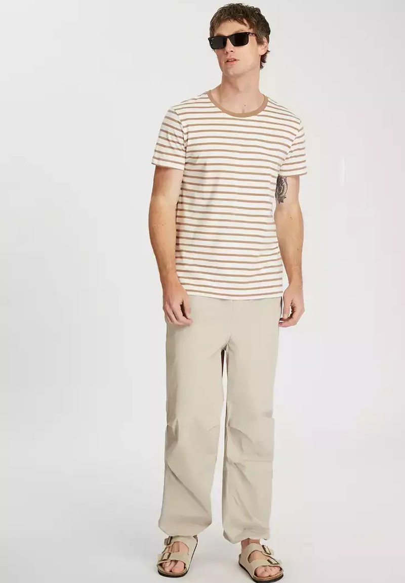 Pasek koszula z krótkim rękawem w kolorze beżowym i brązowym, zestawiona z jasnobeżowymi spodniami. Nosi sandały i ciemne okulary przeciwsłoneczne. Widać tatuaż na ramieniu.