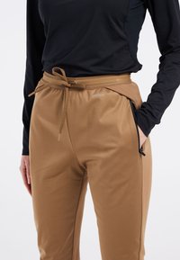 Bruine sportbroek met een trekkoord in de taille, zijzakken met rits en een gladde textuur. Gepaard met een zwart overhemd met lange mouwen.