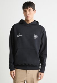 Hoodie noir en tissu à texture lisse. Présente des motifs graphiques blancs, une poche avant et une capuche avec cordon de serrage.