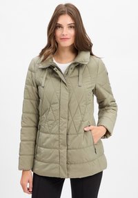 Junge Frau trägt eine hellolivgrüne Steppjacke mit Reißverschluss und Druckknöpfen, mit einer Hand in der Tasche, vor einem weißen Hintergrund stehend.
