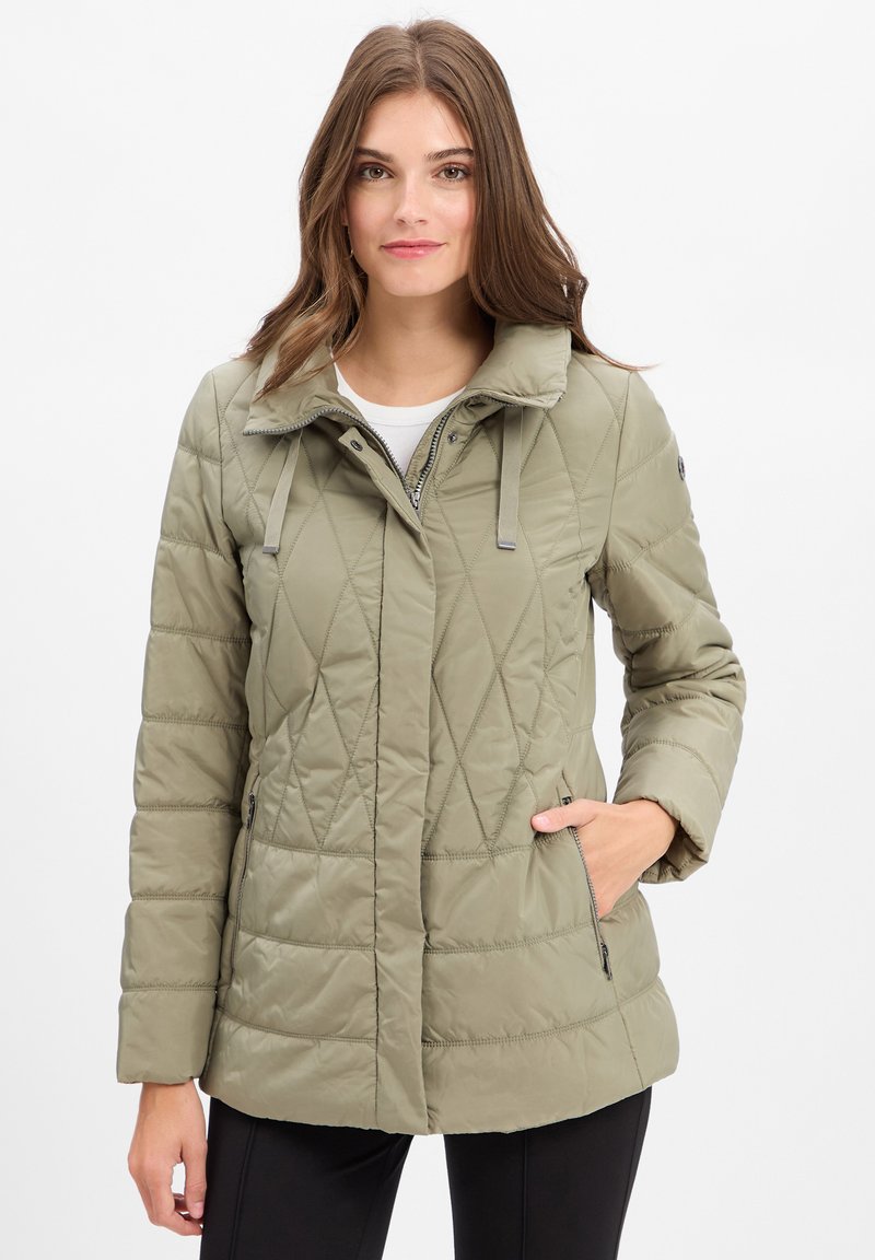 Junge Frau trägt eine hellolivgrüne Steppjacke mit Reißverschluss und Druckknöpfen, mit einer Hand in der Tasche, vor einem weißen Hintergrund stehend.