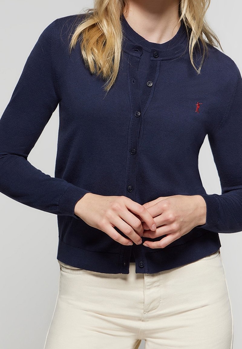 Námořnicky modrý kardigan z měkkého pleteného materiálu s kulatým výstřihem, zapínáním na knoflíky a vyšitým logem na hrudi. Ruce jsou umístěny vpředu.