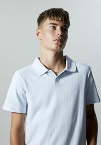 Ljusblå ribbad polo skjorta med spetsig krage och korta ärmar, tillverkad av ett strukturerat tyg, har en enskild knappslå vid halsen.